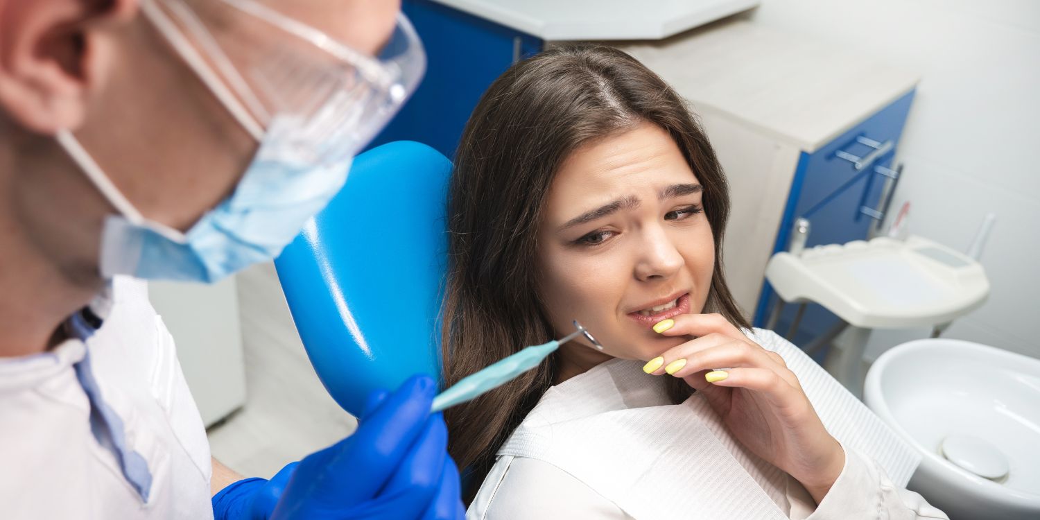 Fiatal nő ijedt arccal ül fogorvosi székben, fogorvos előtérben kezében fogászati szerszámmal.